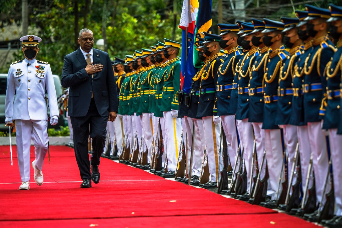 Two men, one in military dress and the other in business attire, walk past a line of people in military dress.
