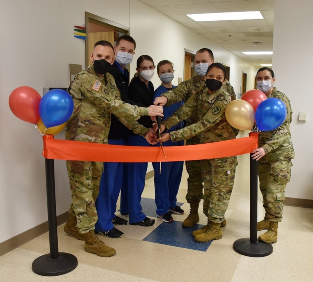 Doctors and nurses are behind a large ribbon with balloons as decoration. They hold large scissors and stand ready to cut the ribbon.