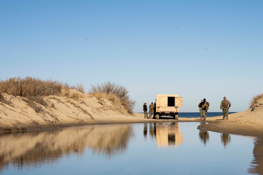Sailors are shown near a military vehicle on the beach, facing the open water.