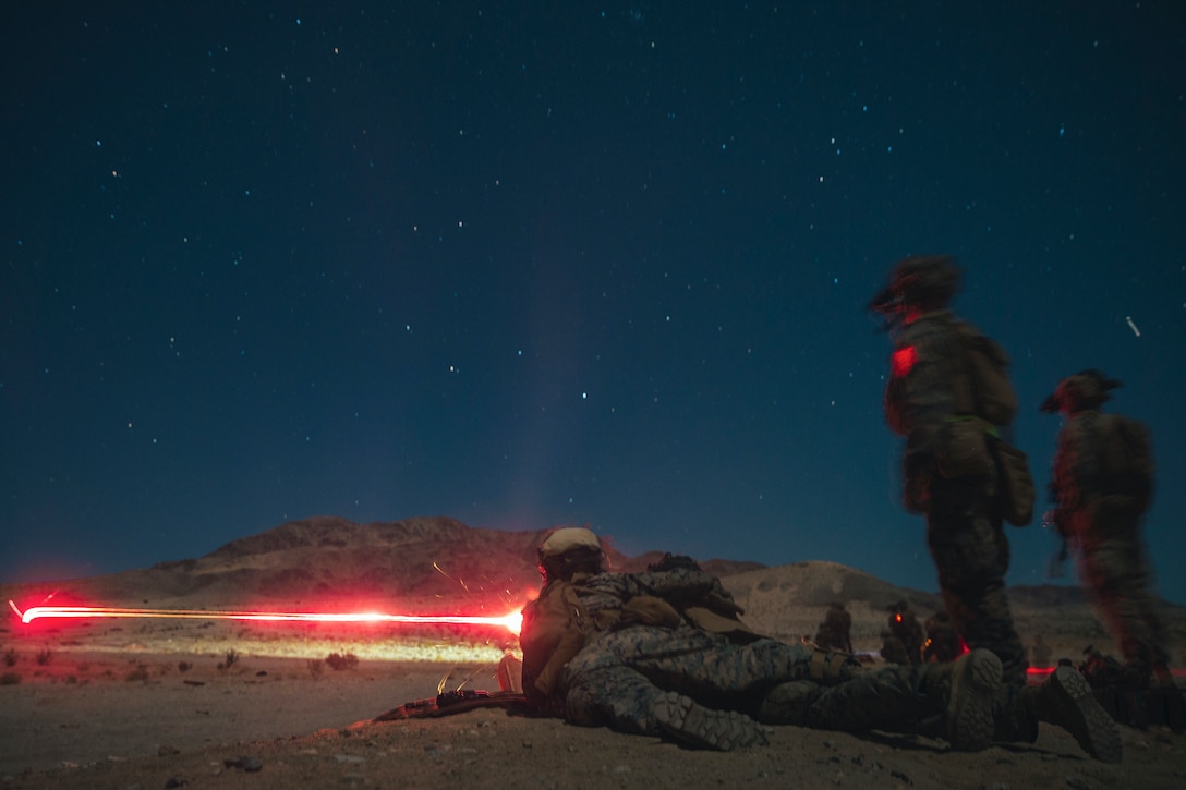 Marines laying on the ground fire a weapon under starry sky illuminated by red light.