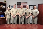 Five naval officers in khaki uniforms standing together in an office with an American flag, photos and plaques in the background.