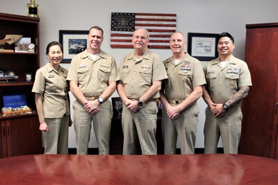 Five naval officers in khaki uniforms standing together in an office with an American flag, photos and plaques in the background.