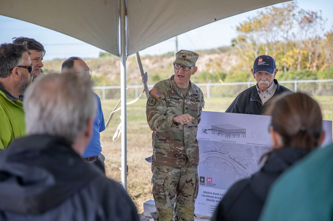 one person in military uniform speaks to several people while pointing to a project design poster