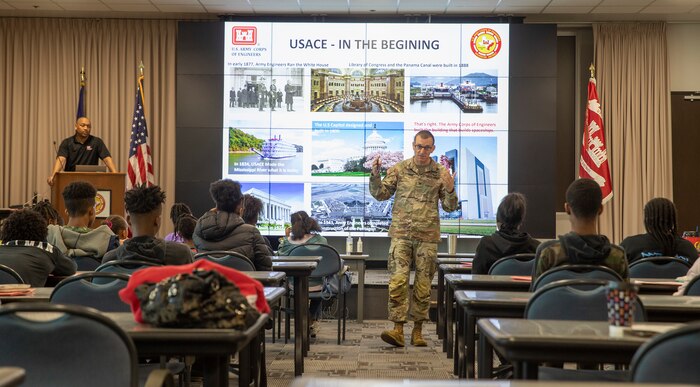 a man in a military uniform stands in front a large presentation screen while talking to seated children