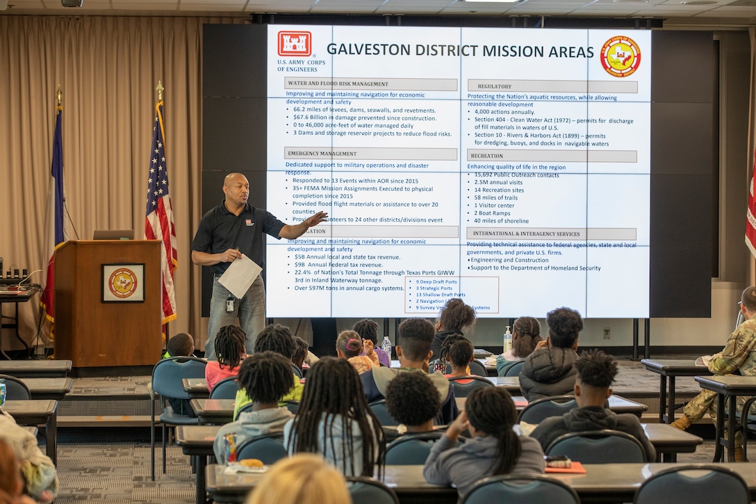 a man stands in front a large presentation screen while talking to seated children