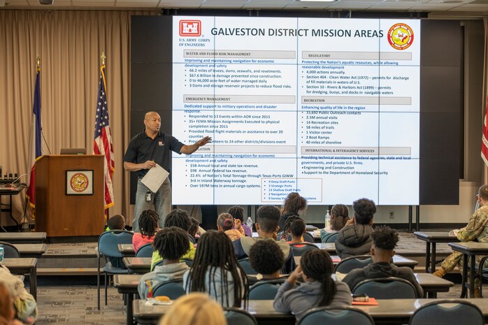 a man stands in front a large presentation screen while talking to seated children