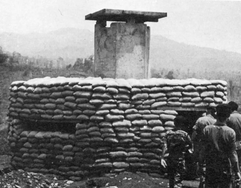 Men walk past a large bunker made of sandbags. A cement tower protrudes from the middle.