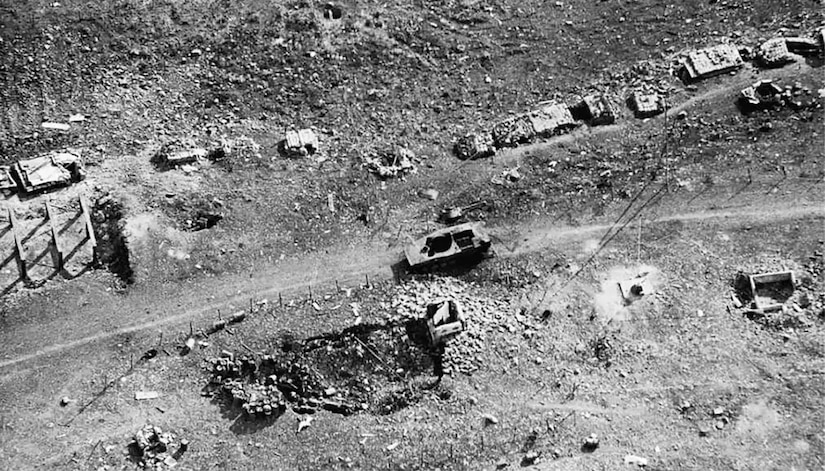 An aerial view of war destruction shows a tank, a destroyed bunker and several small buildings along a dirt path.