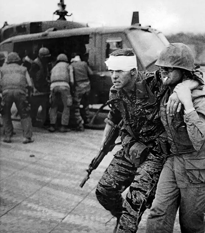 A man with a bandaged head is assisted with walking by  another man. In the background, a person is being helped into a helicopter.