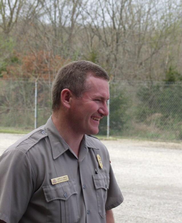 A man in a park ranger uniform is shown in profile with trees in the background.