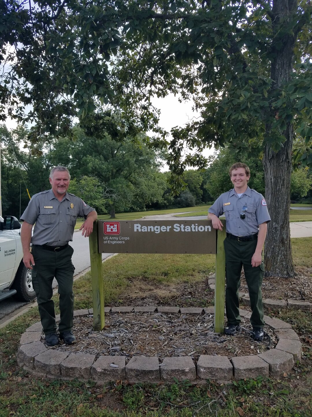 Two men in park ranger uniforms pose on either side of a ranger station sign with trees in the background.