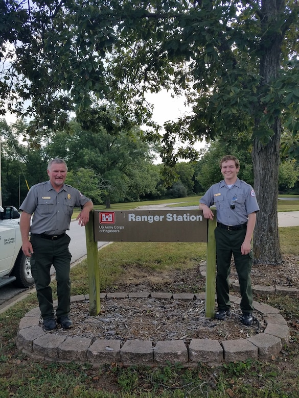 Two men in park ranger uniforms pose on either side of a ranger station sign with trees in the background.
