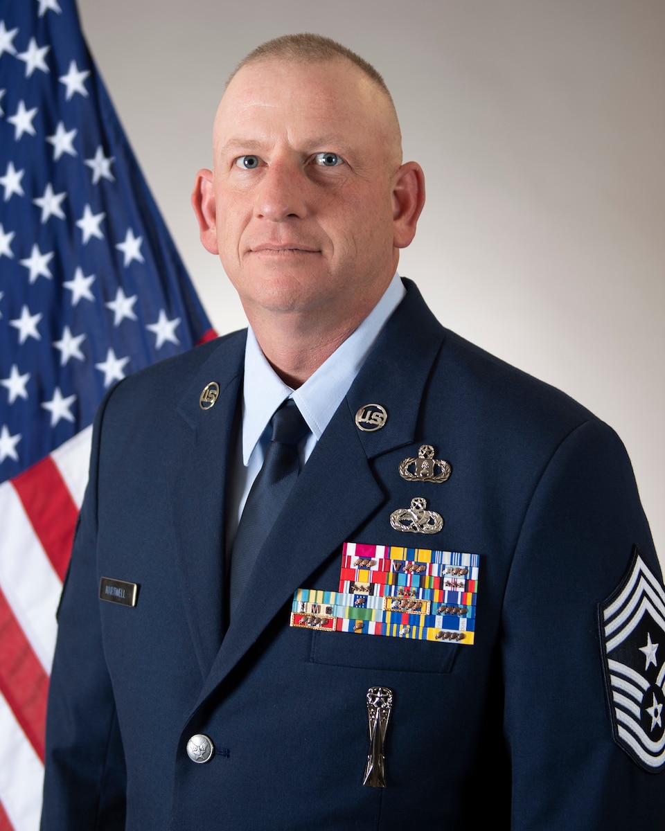A Caucasian male poses for a portrait in front of an American flag with a gray background. The individual is where a U.S. Air Force service blue uniform with a dark blue jacket, light blue shirt and tie. On the left side of the uniform is a silver name tag with the last name of Hartwell. On the right side of the uniform are two silver occupational badges, a large ribbon rack, and a silver missiler badge. A chief master sgt rank is visible on the uniform sleeve.