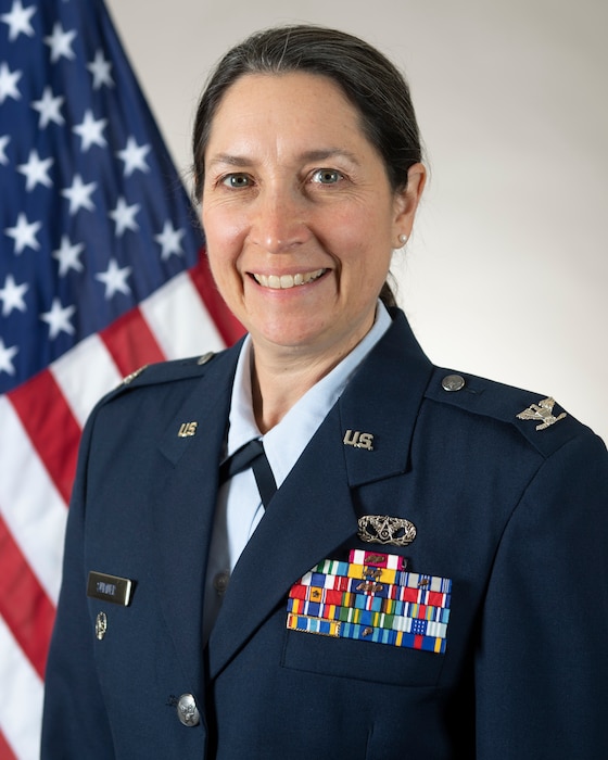 A female U.S. Air Force service member wearing a service dress uniform poses in front of an American flag for a portrait. On the uniform shoulders there are silver colonel ranks, on the left side of the uniform there is a silver nametag that says Sumner on it with a silver command wreathe under the nametag. On the right side of the uniform is a small silver civil engineer occupational badge above a ribbon rack with military awards on it.