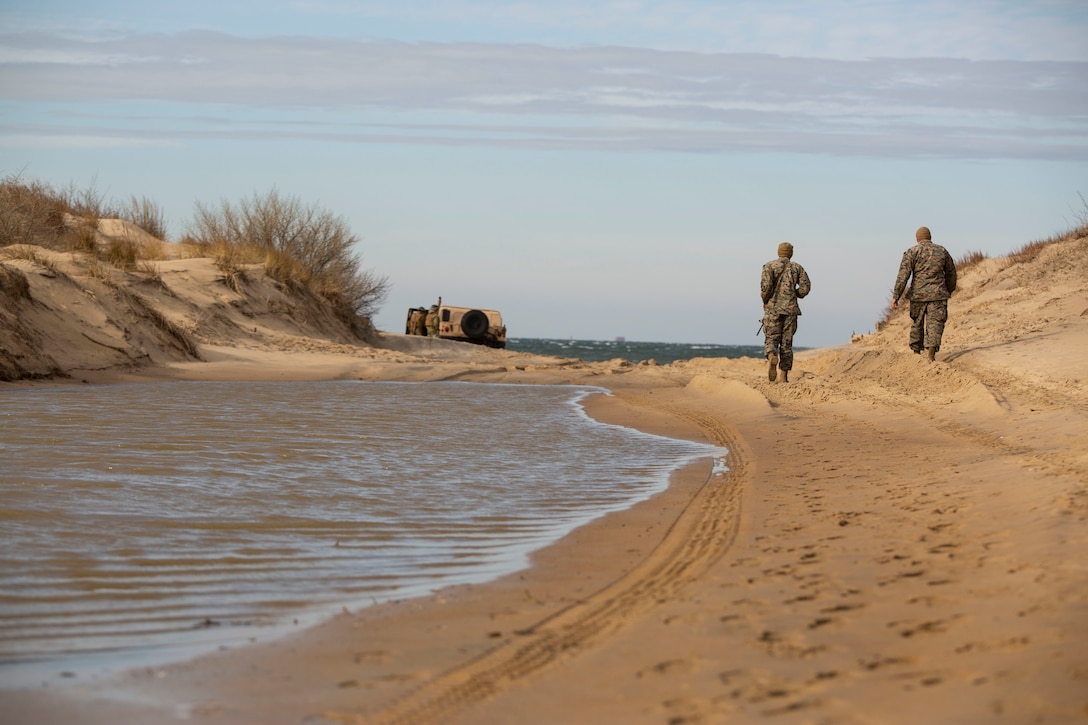 Two service members walk along a beach with a vehicle in the background.