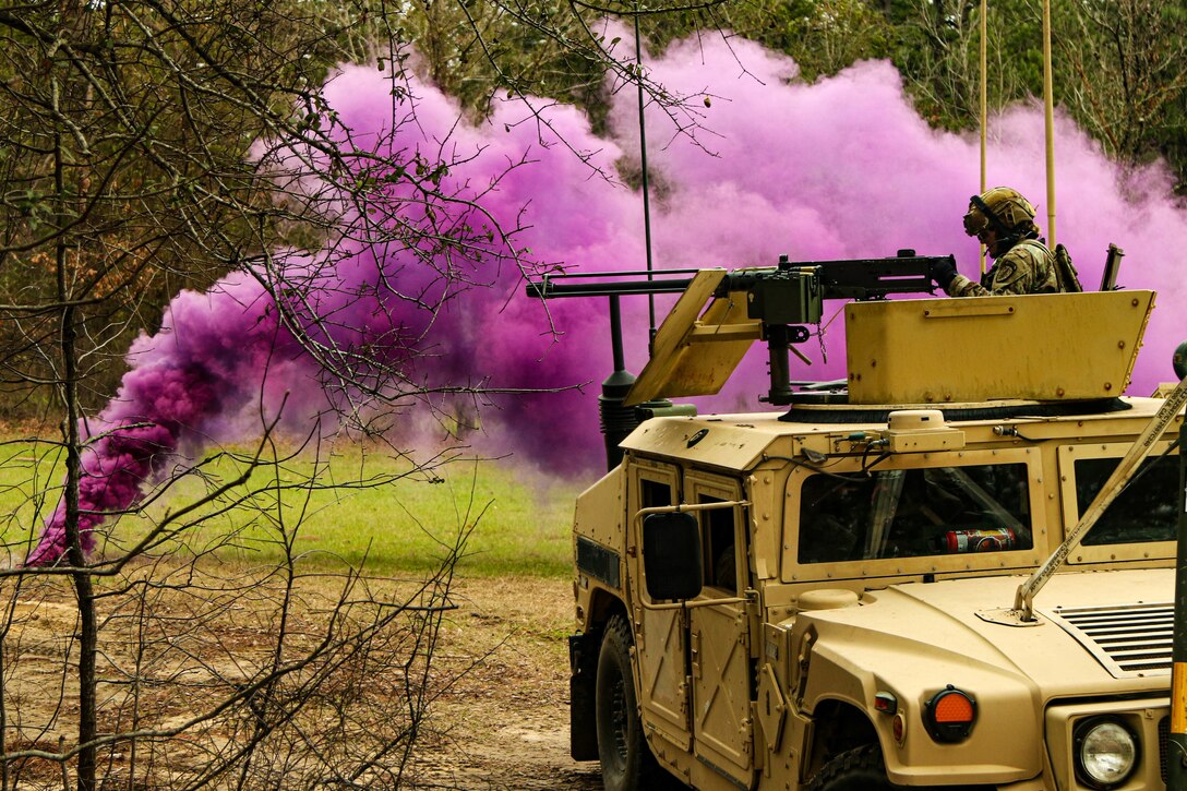 A soldier in a tank participates in an exercise while a canister of purple smoke is released nearby.