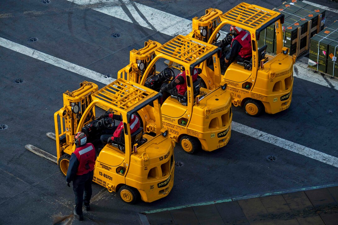 Several sailors sit inside and stand next to three parked yellow forklifts on the deck of a ship.