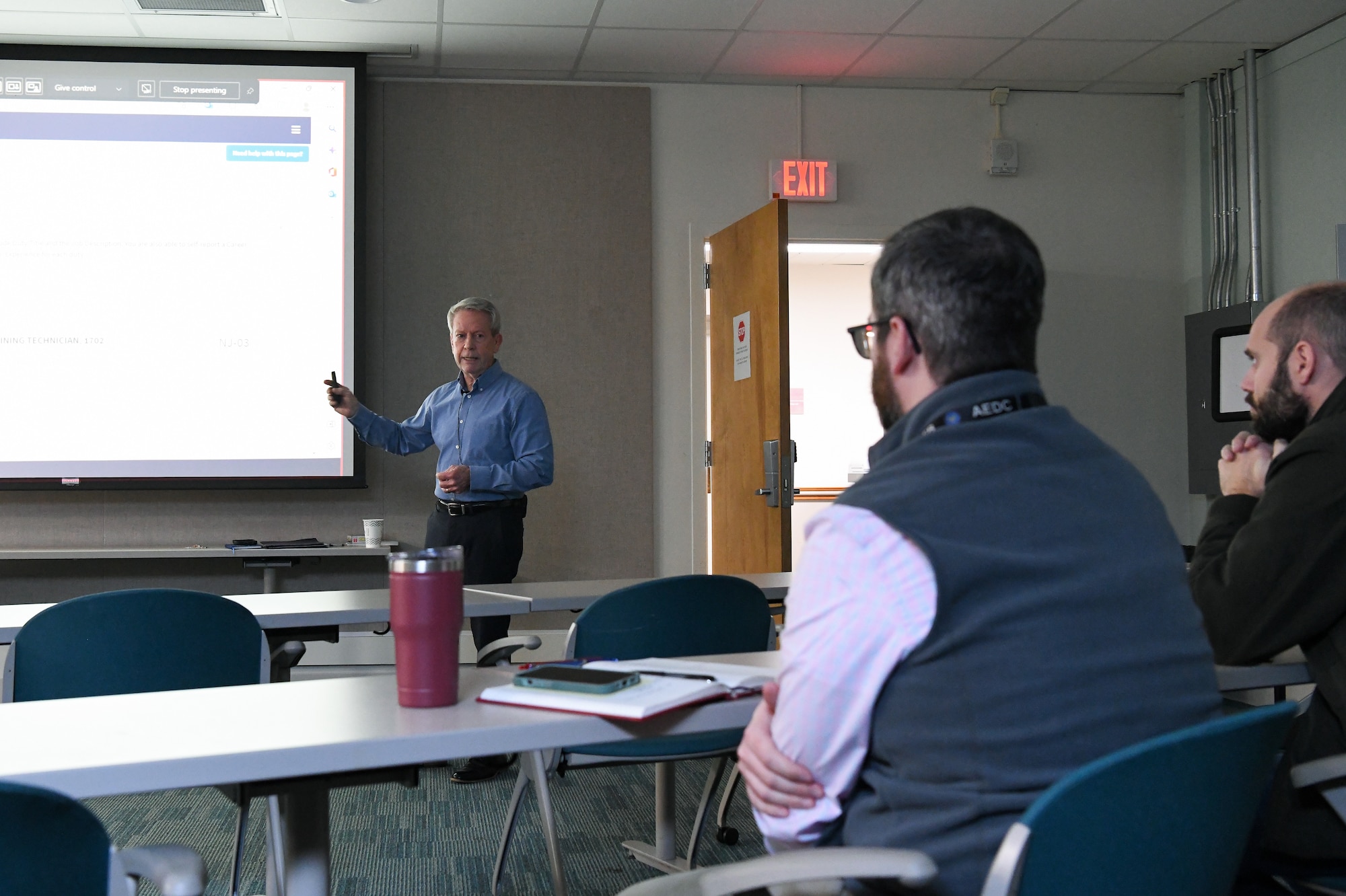 Mike Dent, standing at left, the Chief, Technical Management Branch, Plans and Programs Division, Arnold Engineering Development Complex, gives a presentation. Two men are seated at a table at right.