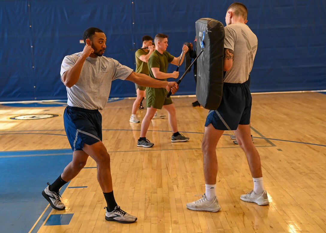 Military members in physical training uniforms strike pads with batons.