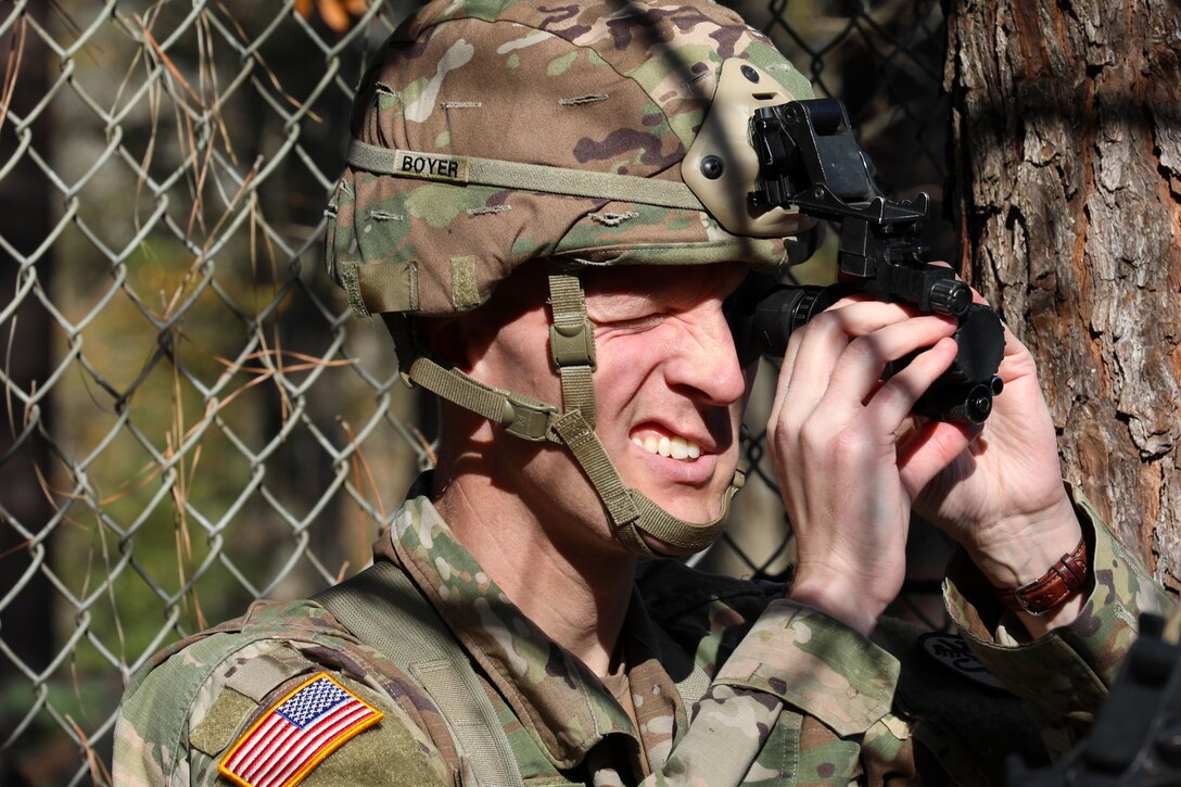 A uniformed service member is shown close up, operating a night vision device.