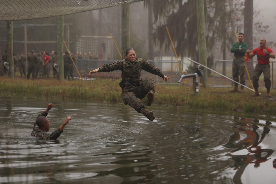 A Marine Corps recruit jumps into small body of water as fellow Marines cheer.