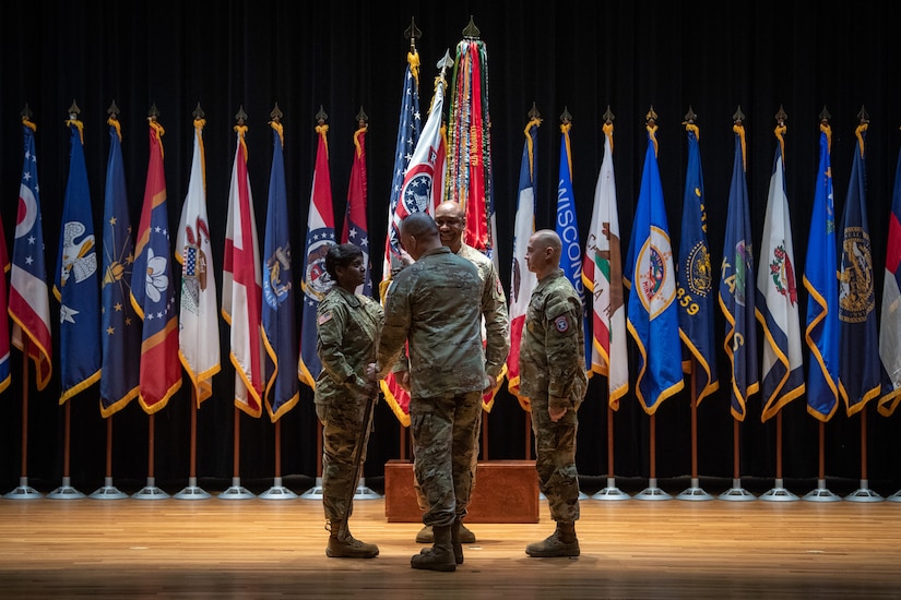 four people wearing u.s. army uniform pass a flag on a pole between them.