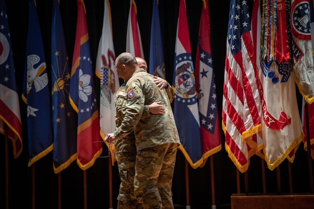 two men hug in front of flags wearing u.s. army uniform.