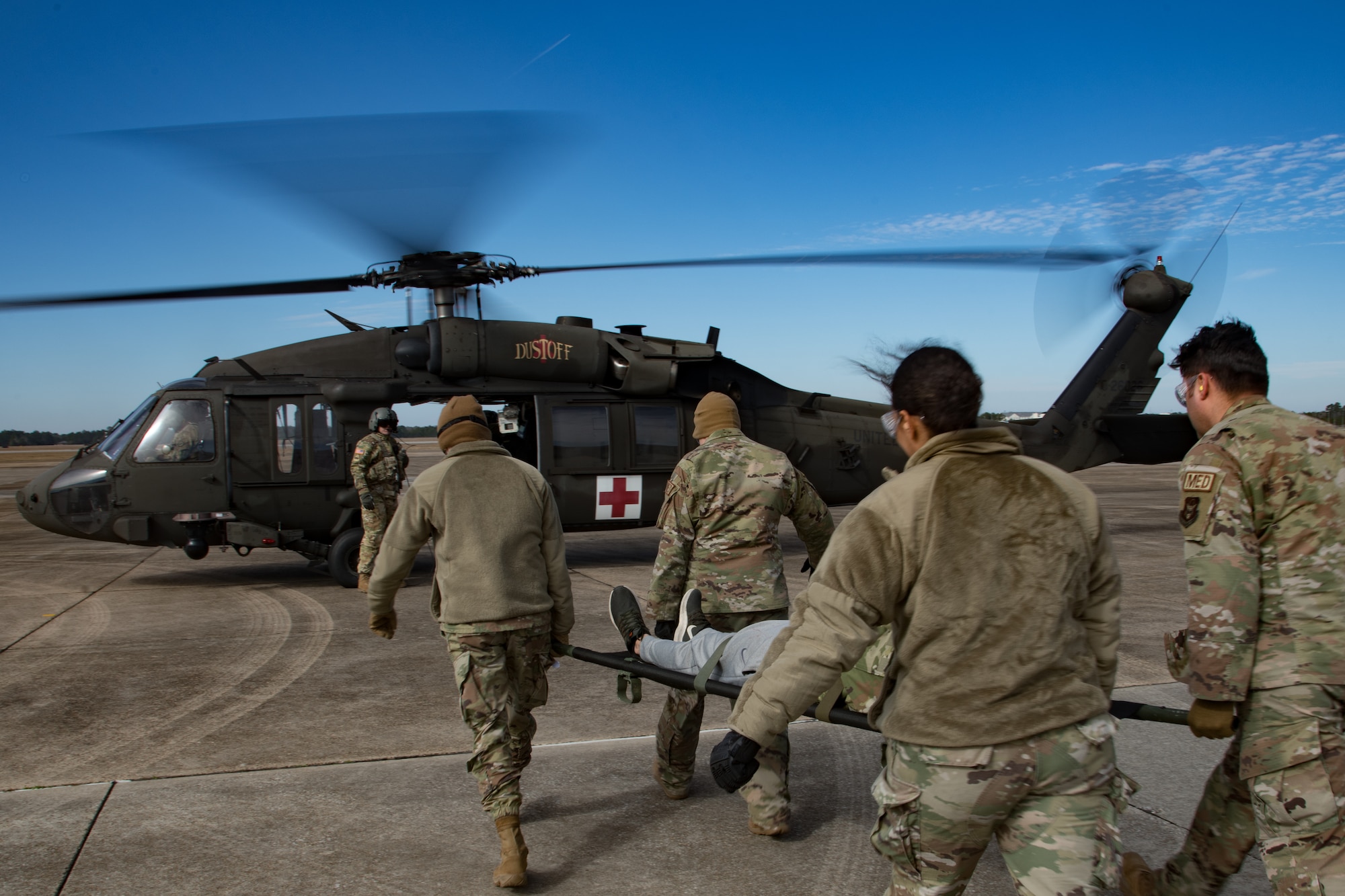 U.S. Airmen in uniform transport a litter to a UH-60 Blackhawk helicopter with its engine running.