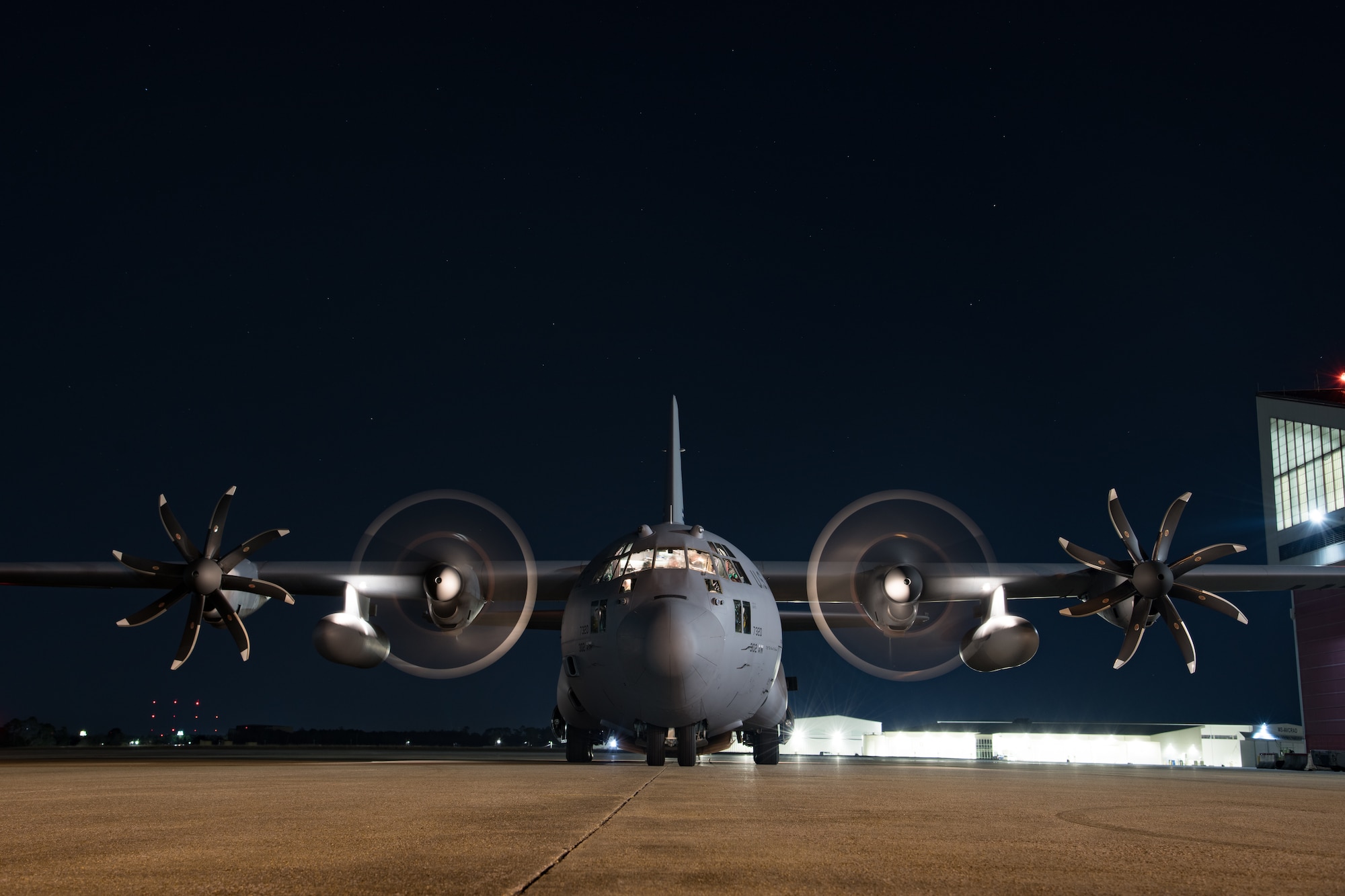 Front-on visual of a C-130H aircraft parked at night with two outboard propellers still and two inboard propellers spinning and the crew compartment light on.