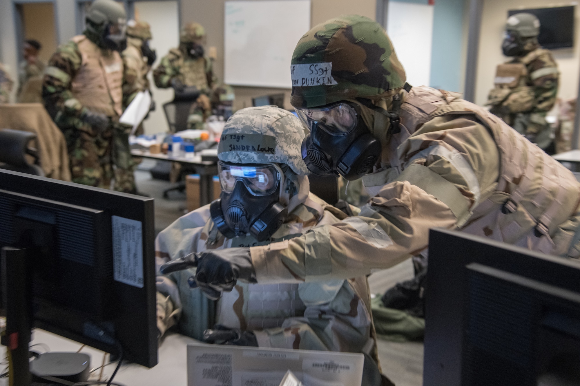 Two Airmen in chemical protective equipment look at a computer screen reflected in their gas mask visor.