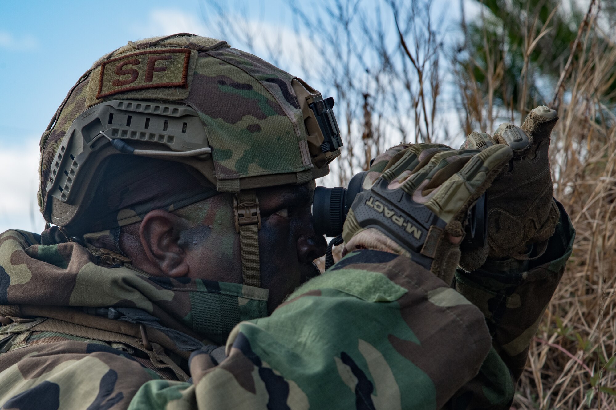 An Airman wearing chemical protective gear and a helmet looks through a pair of binoculars.