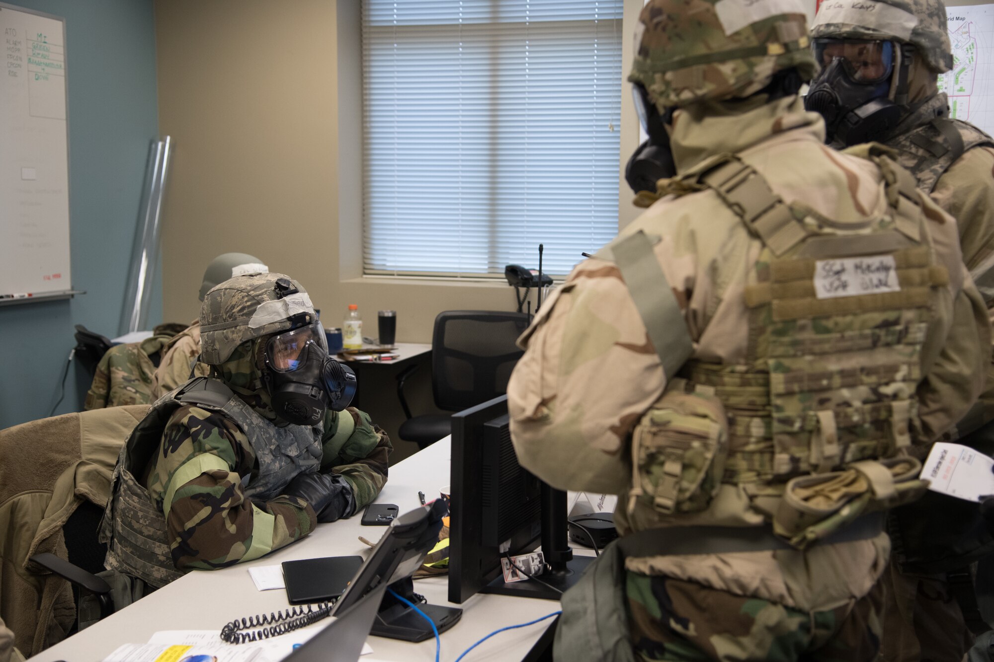 Two Airmen in chemical protective gear report to the wing commander sitting at a desk during a training exercise.