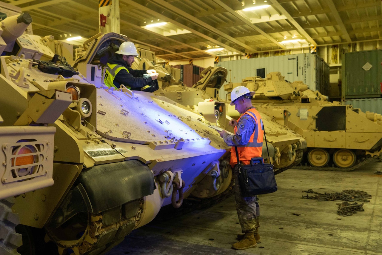 A soldier checks paperwork while another soldier stands in a tank.