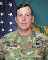 Man in U.S. Army uniform standing in front of two flags.