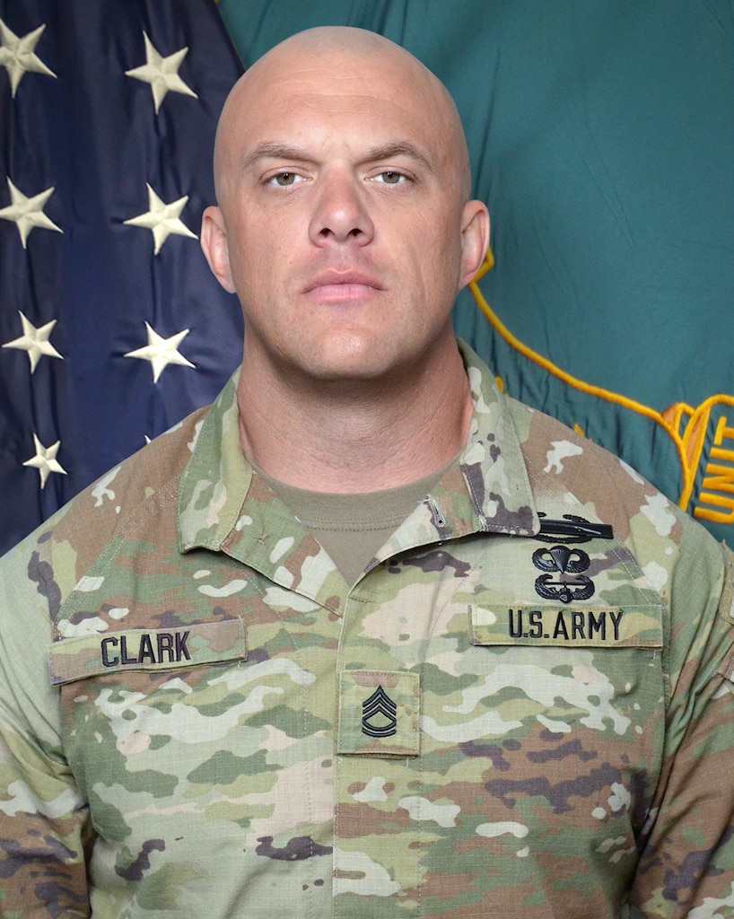 Man in U.S. Army uniform standing in front of two flags.
