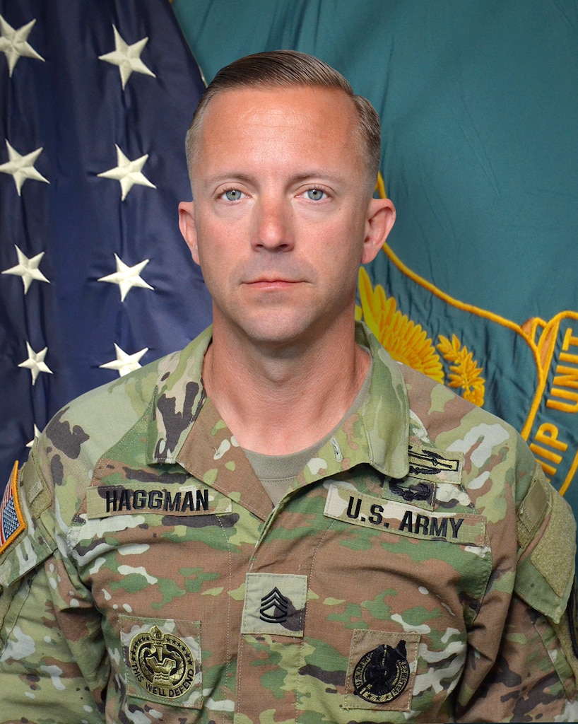 Man in U.S. Army uniform standing in front of two flags.