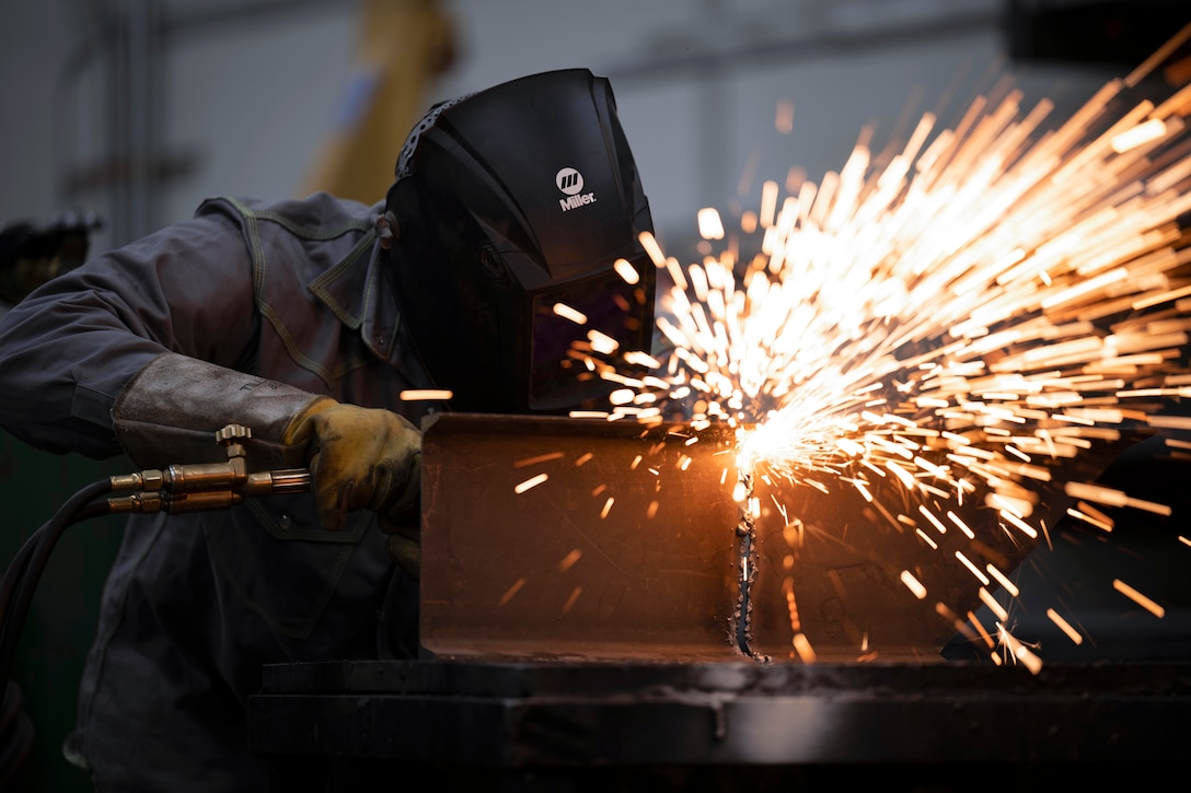 Sparks fly as an airman wearing protective gear welds a piece of metal.