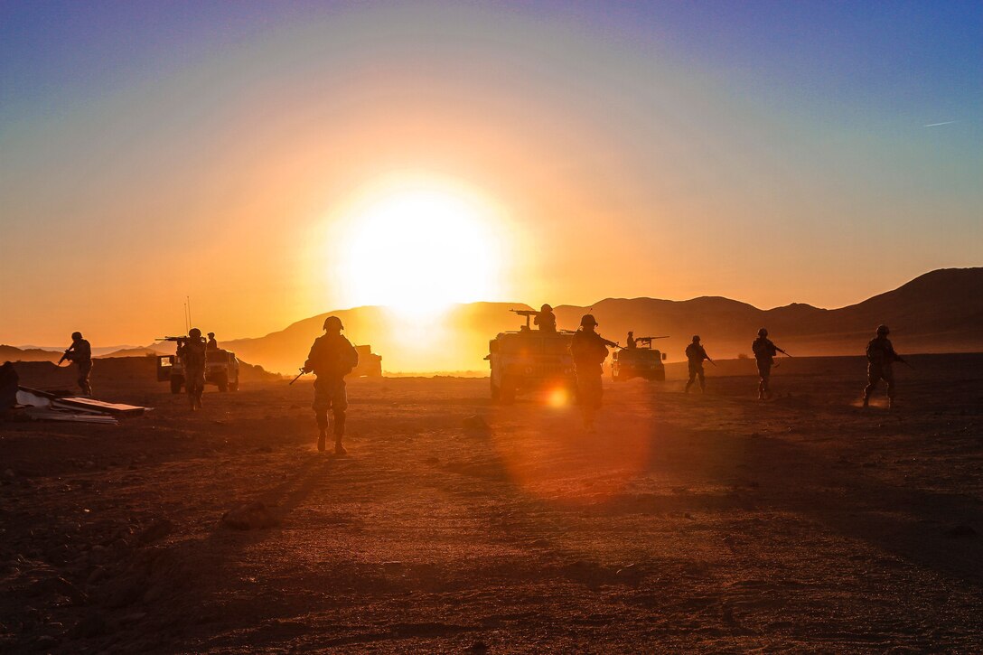 Soldiers shown in silhouette walks through desert terrain as the sun shines behind.
