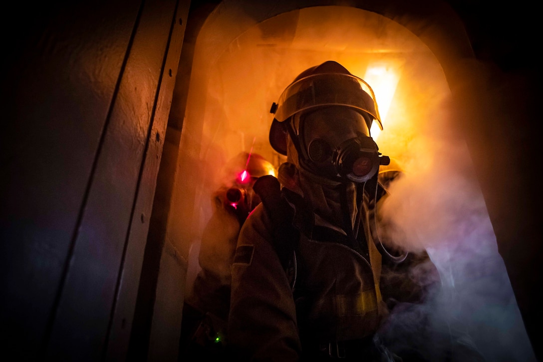 A sailor wearing fire protection gear walks though a smoky doorway.