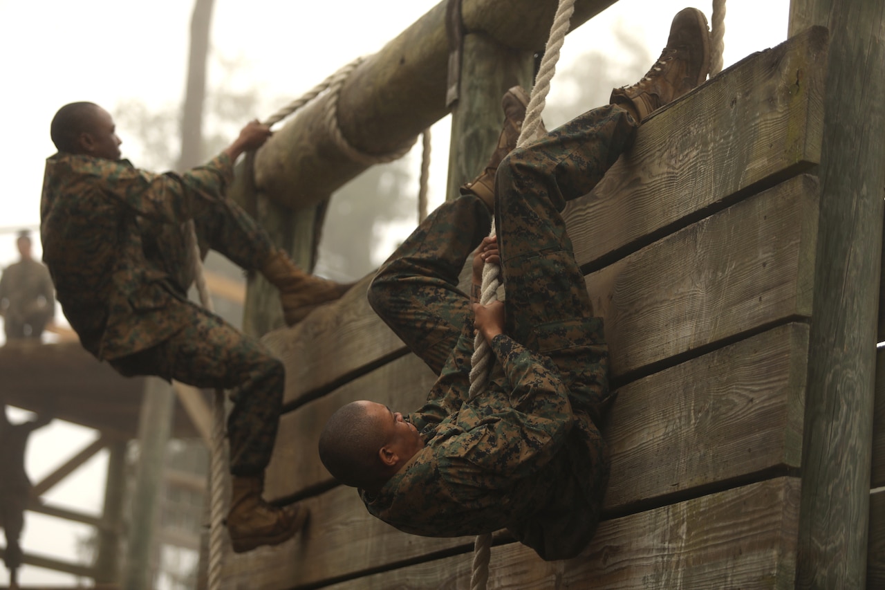 Marine Corps recruits use ropes to climb up a wooden wall.