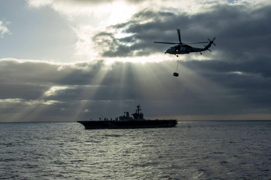 A military helicopter prepares to make an ammunition delivery while flying over a Navy ship at sea.