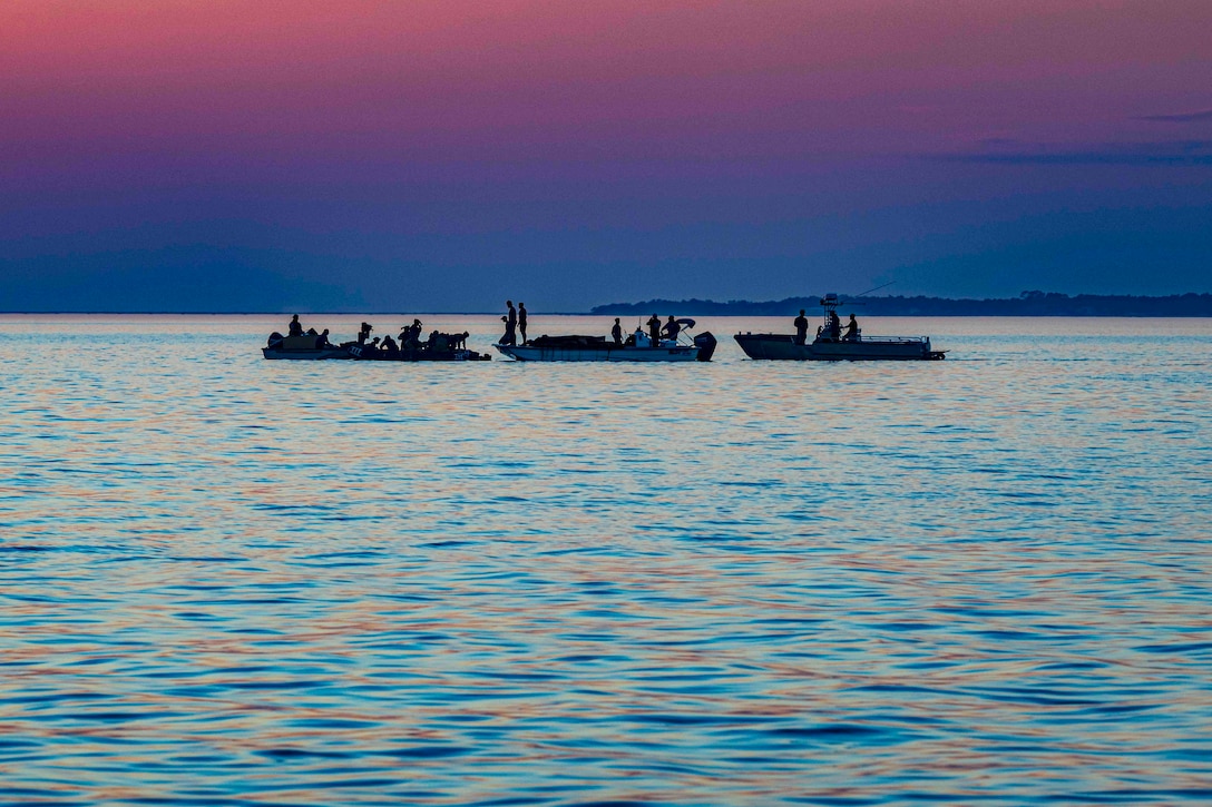 Service members work on three watercraft in water against a purple and pink sky.