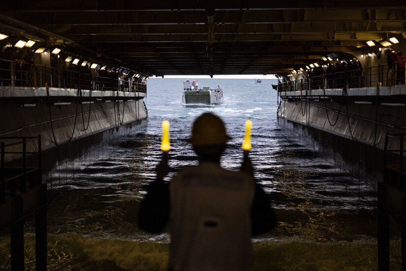 A sailor uses two lights to guide a small boat into the well deck of a ship.