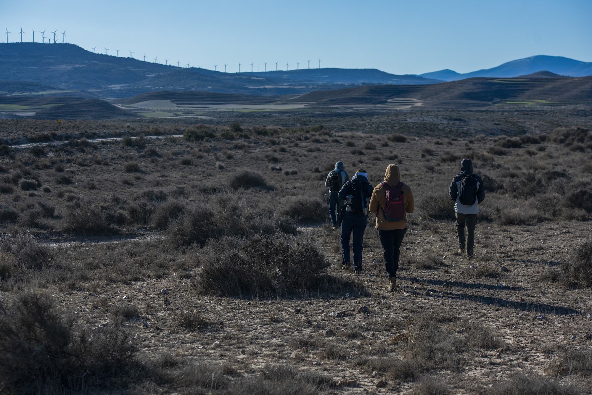 Four people walk through a desert and mountainous region. There are various bushes on the ground. Windmills can be seen on the mountains in the background.