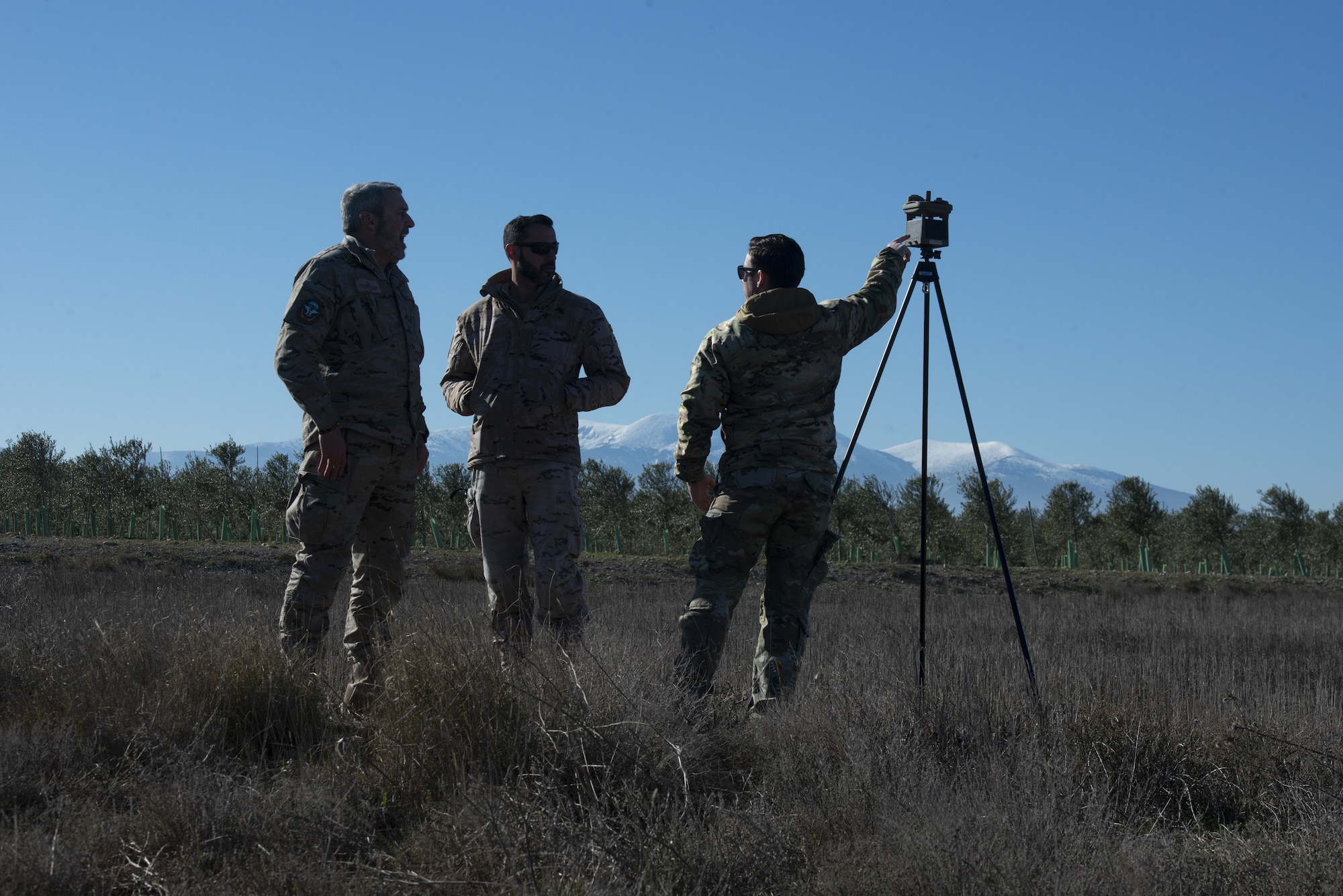 Three people are next to a tripod stand. There is a blue sky and white and grey mountains in the distance