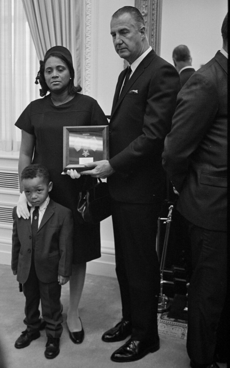 A man holds a small plaque in front of a solemn-looking woman and a young boy.