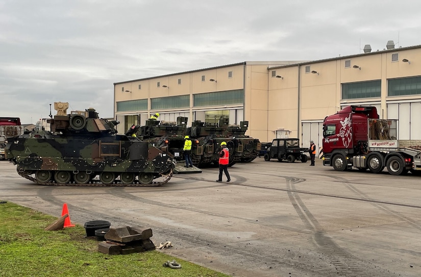 People walk around three tanks and two tractor-trailers that are parked outside a large building.