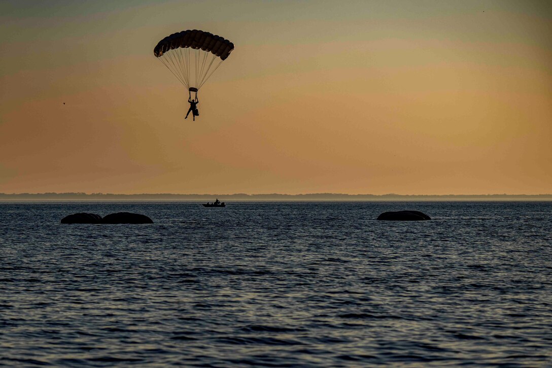 Sailor shown in silhouette parachutes to water where empty uninflated boats sit.