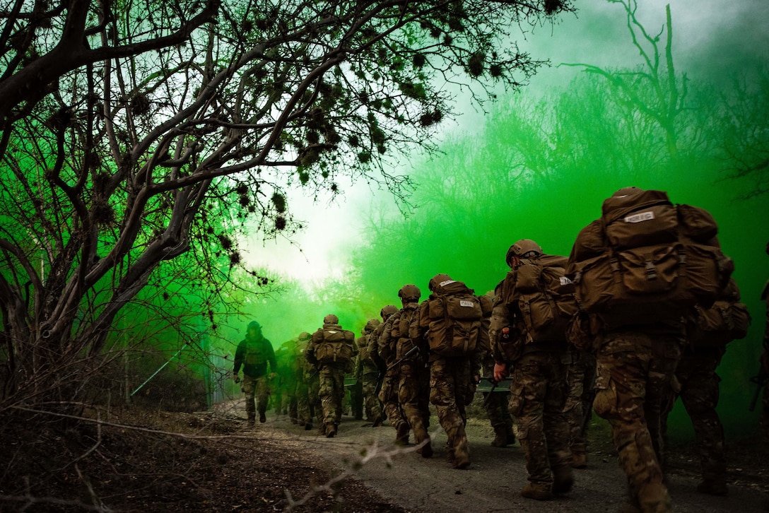 Airmen walk uphill in a wooded area, surrounded by green smoke.