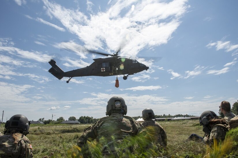 Soldiers kneel in the grass as a helicopter flies above.